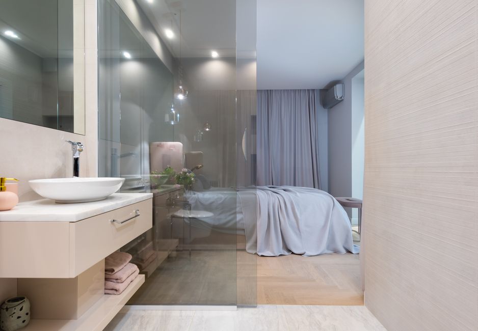 Interior of light modern restroom with sink and faucet on cabinet under mirror on wall next to glass door to bedroom with bed